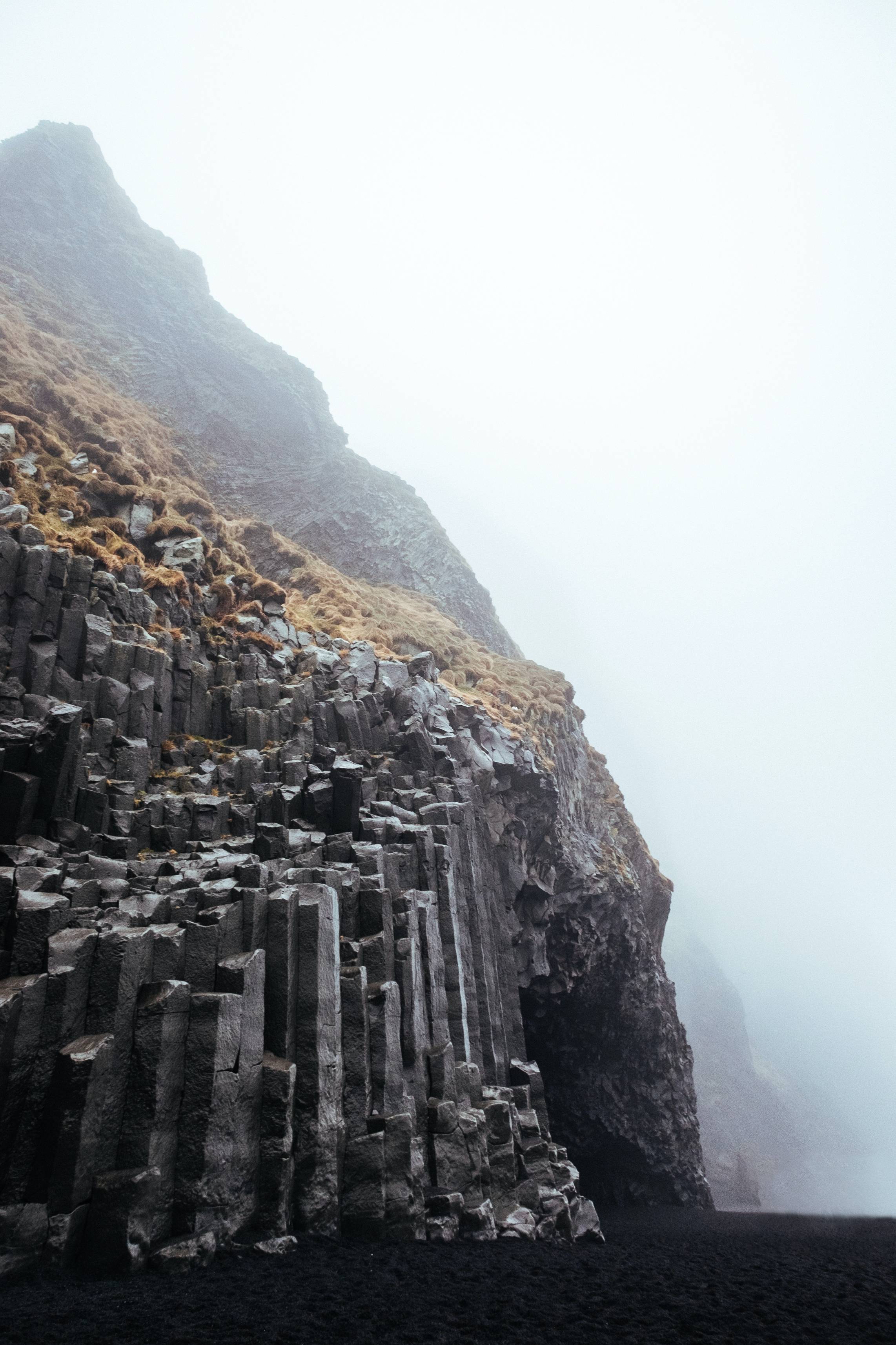 Reynisfjara-basalt-columns-and-cave-opening-seen-from-beach-with-yellow-grass-on-top-surrounded-by-the-ocean-mist
