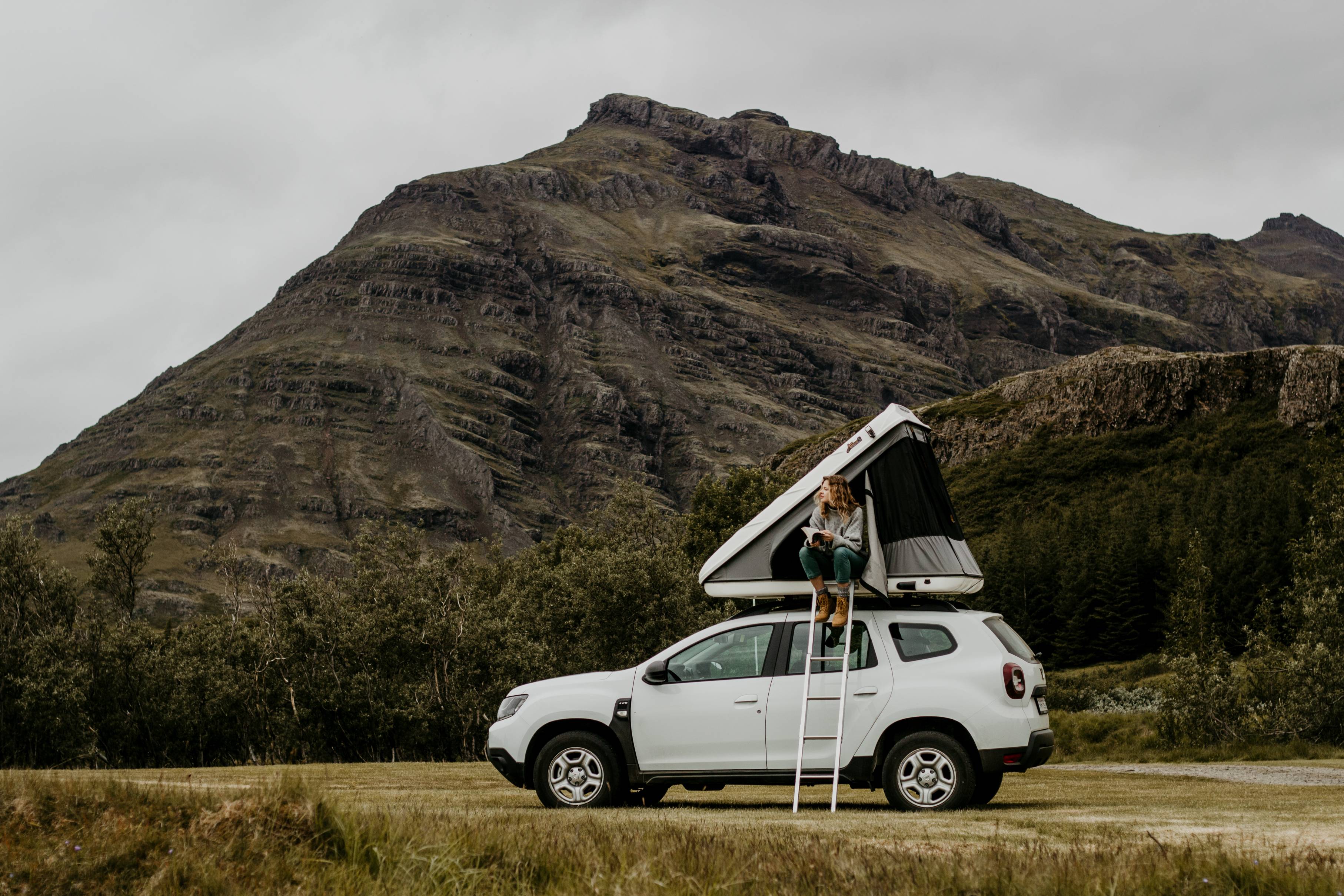 Woman-with-curly-hair-in-a-roof-tent-on-a-white-Dacia-Duster-4x4-in-Iceland-reading-a-book-in-summer
