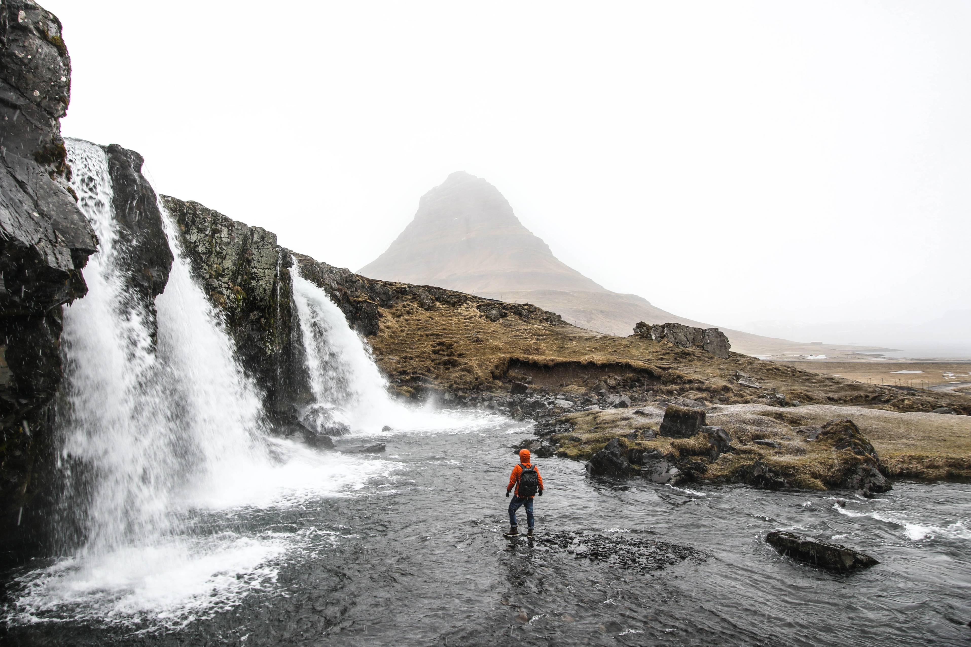 man-in-a-orange-jacket-walking-in-stream-on-a-cold-cloudy-day-in-iceland-with-mount-kirkjufell-in-background